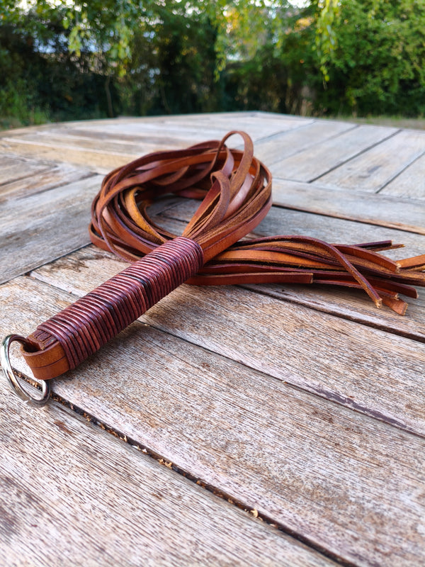 Flogger lourd AdryX en lanières de cuir épais 550 g, manche ligaturé et anneau, posé sur une table en bois en extérieur