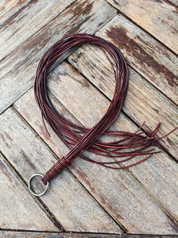 Flogger en cordons de cuir AdryX posé en boucle sur une table en bois, anneau métallique pour la prise à deux doigts