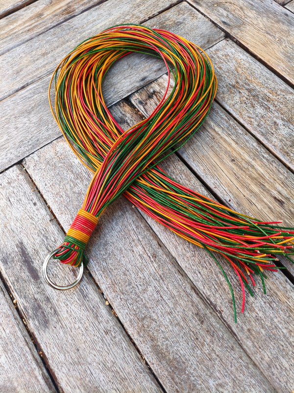 Martinet Rastacouère AdryX en cordons de cuir rouge, jaune et vert, 100 cm, posé en boucle sur une table en bois