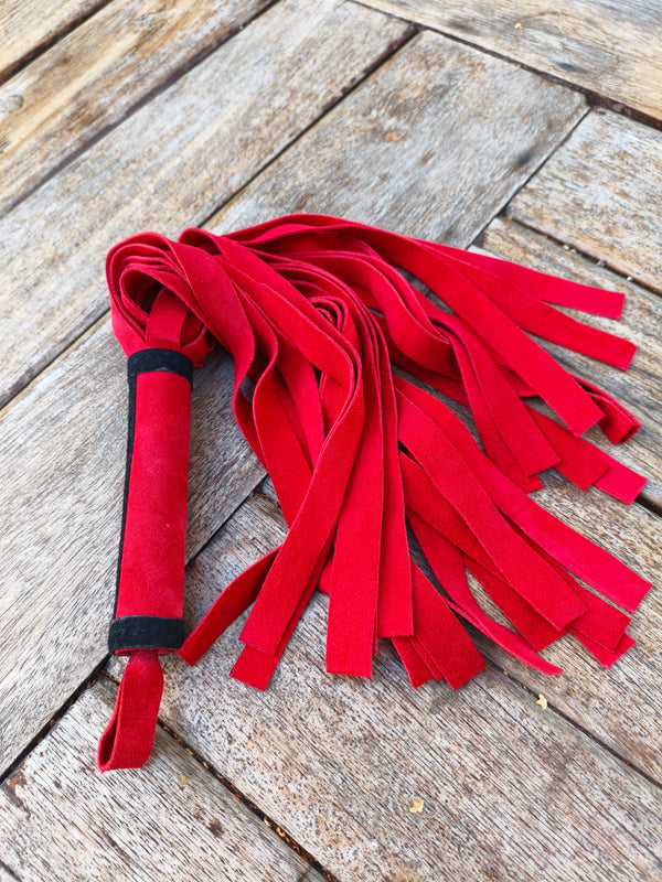 Martinet doux AdryX en croûte de cuir rouge, 40 cm, 27 lanières et poignée rouge à bande noire, posé sur une table en bois