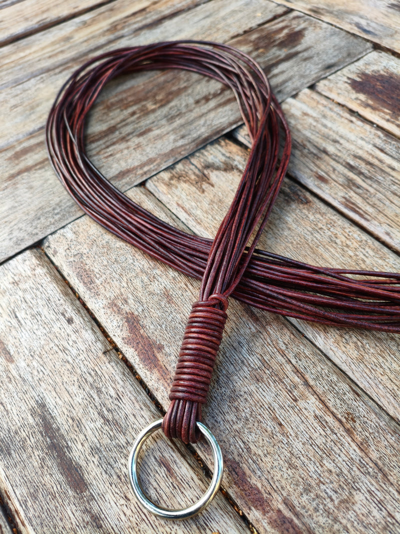 Flogger AdryX en cordons de cuir, 100 cm, cordons repliés en boucle et manche ligaturé au premier plan, sur une table en bois