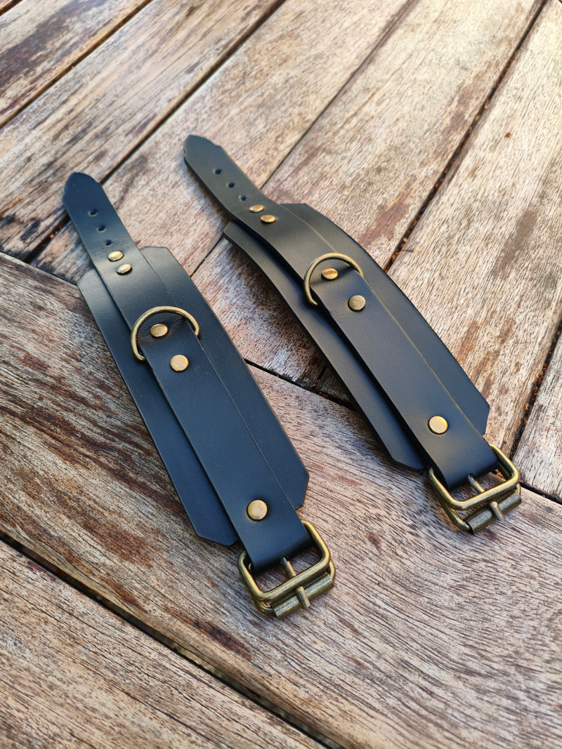 Paire de menottes AdryX en cuir noir à boucles laiton, posées à plat sur une table en bois
